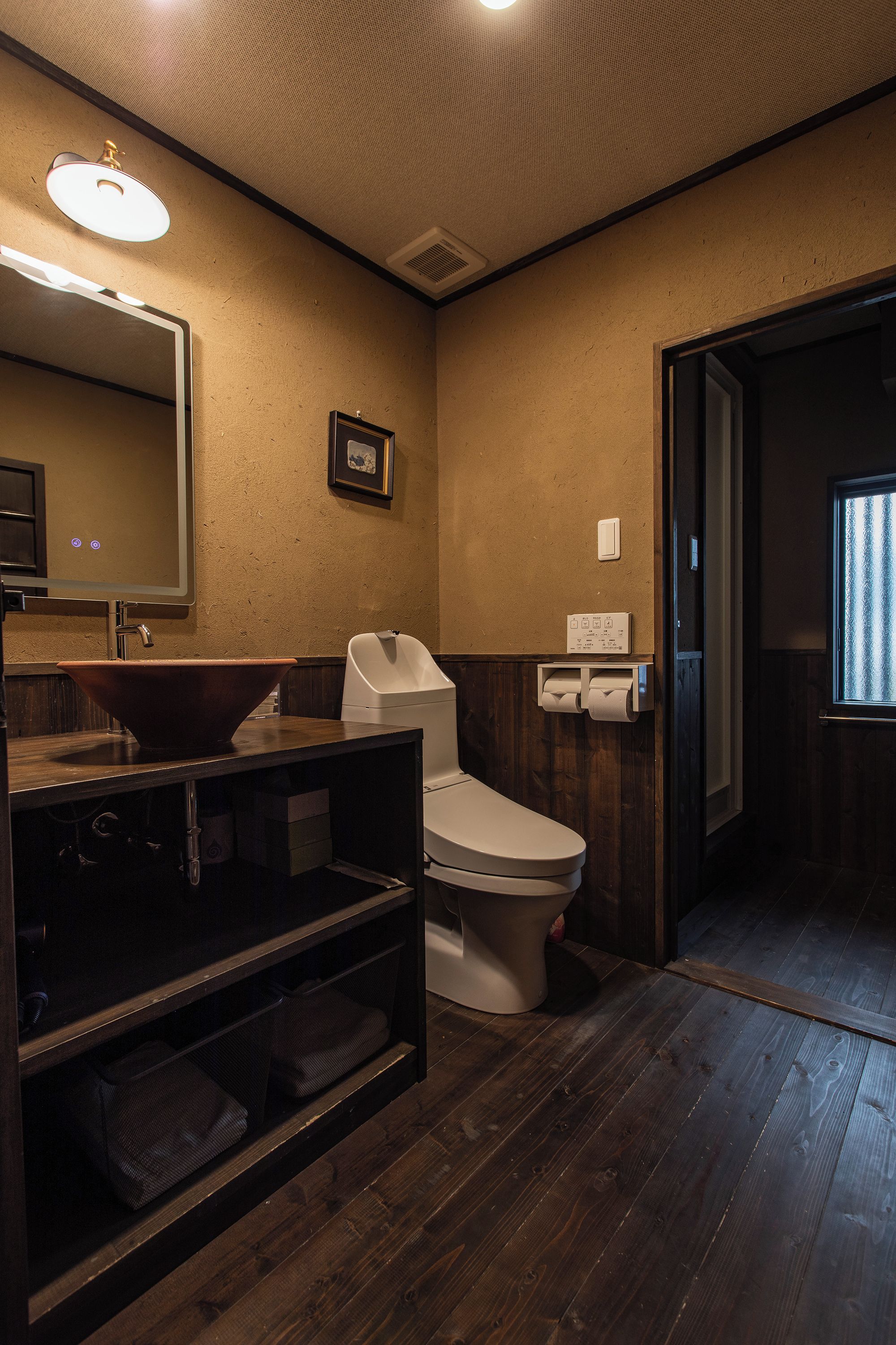 A bathroom with a wooden floor, a sink with a mirror, a toilet, and a framed picture on the wall.