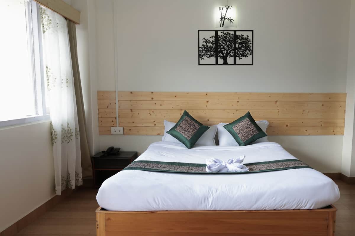 A neatly made bed with white linens and two green patterned pillows, a wooden headboard, a wall-mounted tree decoration, and a bedside table with a phone.