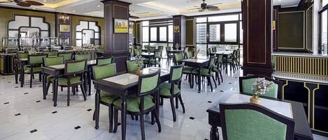 A spacious restaurant with high ceilings, green upholstered chairs, and large windows offering city views.
