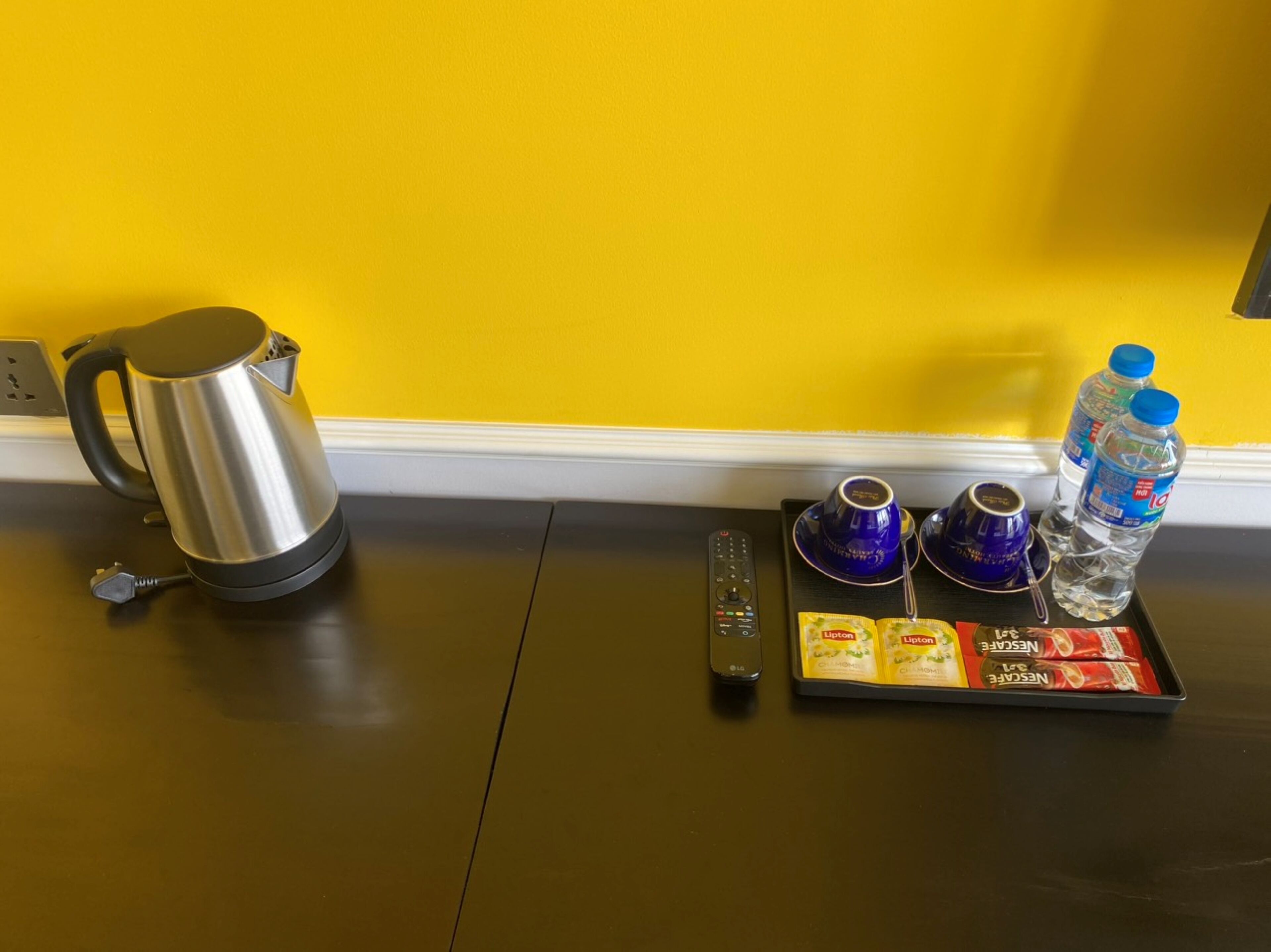 A kitchen area with a kettle, a remote control, and a tray with Lipton tea and water bottles.