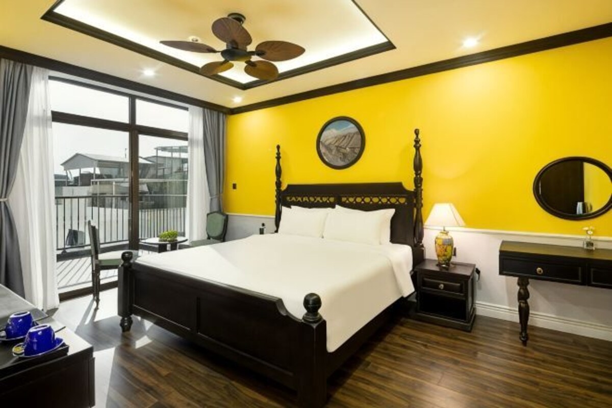 A hotel room with a large bed, a ceiling fan, a window with curtains, a bedside table with a lamp, and a mirror on the wall.