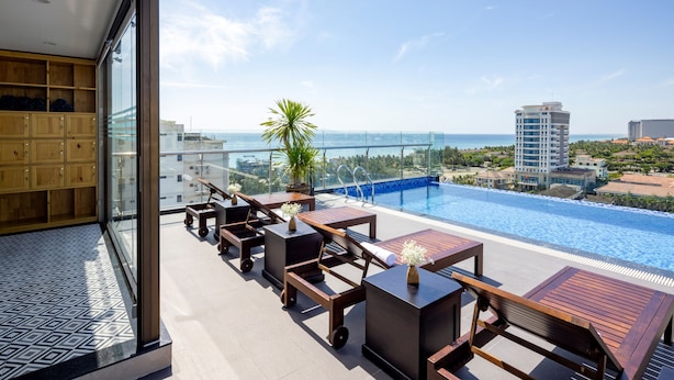 A rooftop pool area with lounge chairs and tables, a clear view of the ocean, and a modern building in the background. - Charming Beauty Hotel (Da Nang)