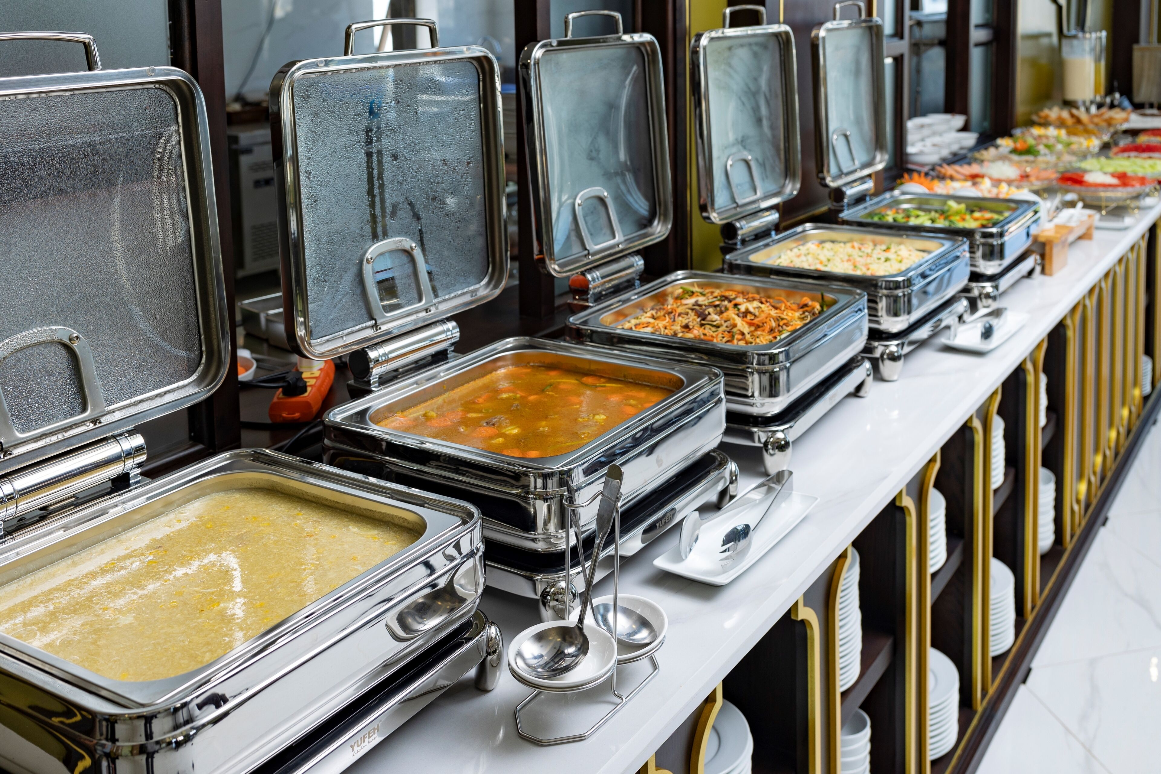A buffet line with multiple self-service food stations, each with a covered serving tray and a spoon.