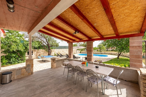 An outdoor patio with a table and chairs, a pool in the background, and stone walls.