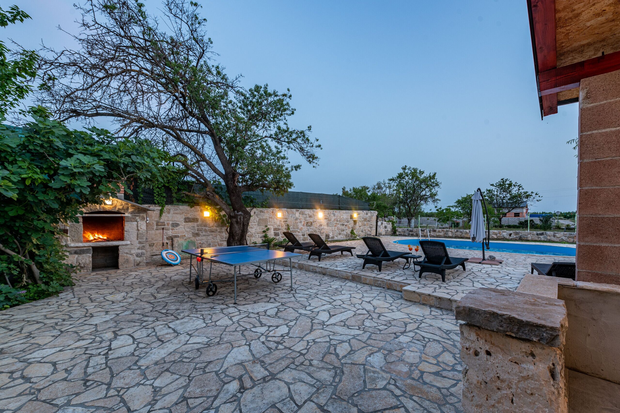 Un patio exterior con suelo de piedra, un pozo de fuego, sillones de descanso y una piscina.