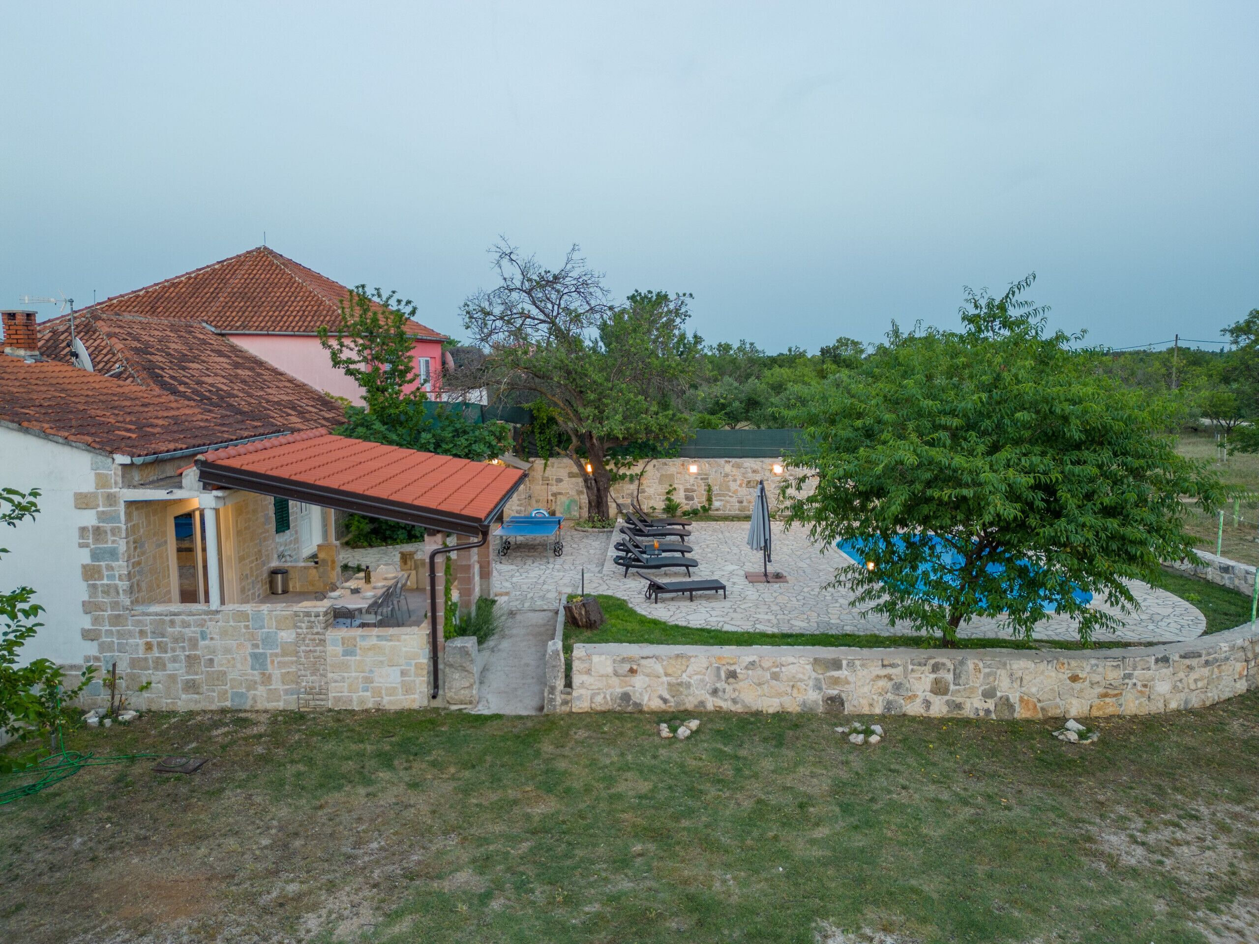 A stone house with a tiled roof, a swimming pool, and a patio area with lounge chairs and a barbecue.
