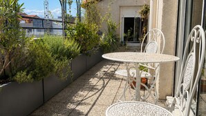 Ein Balkon mit zwei weißen Metalltischen und Stühlen, umgeben von Blumentöpfen und Blick auf ein Gebäude.
