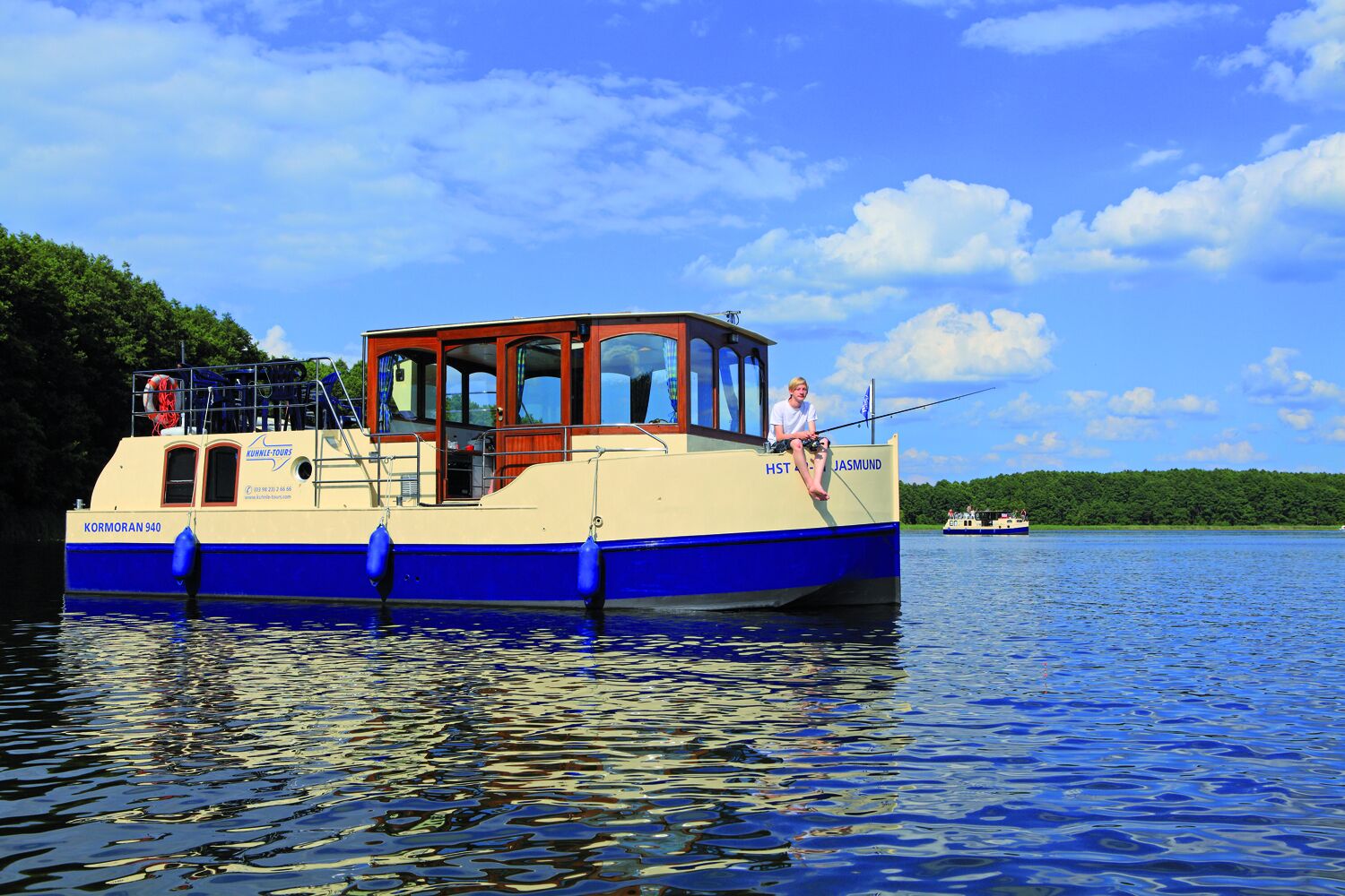 A boat named 'Kormoran 940' with a cabin and a person fishing, surrounded by calm water and greenery.