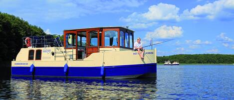 A boat named 'Kormoran 940' with a cabin and a person fishing, surrounded by calm water and greenery.