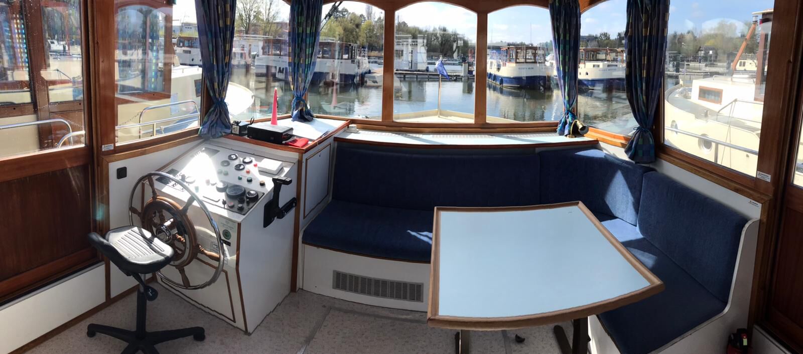 Een bootcabine met een bedieningspaneel, een stuurwiel, een beklede zithoek en uitzicht op andere boten door grote ramen.
