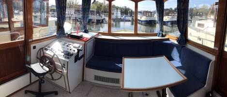 Een bootcabine met een bedieningspaneel, een stuurwiel, een beklede zithoek en uitzicht op andere boten door grote ramen.