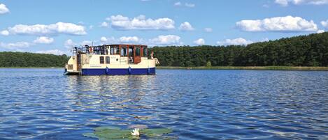 A boat with a cabin and deck seating, floating on calm water with a forested shoreline in the background.