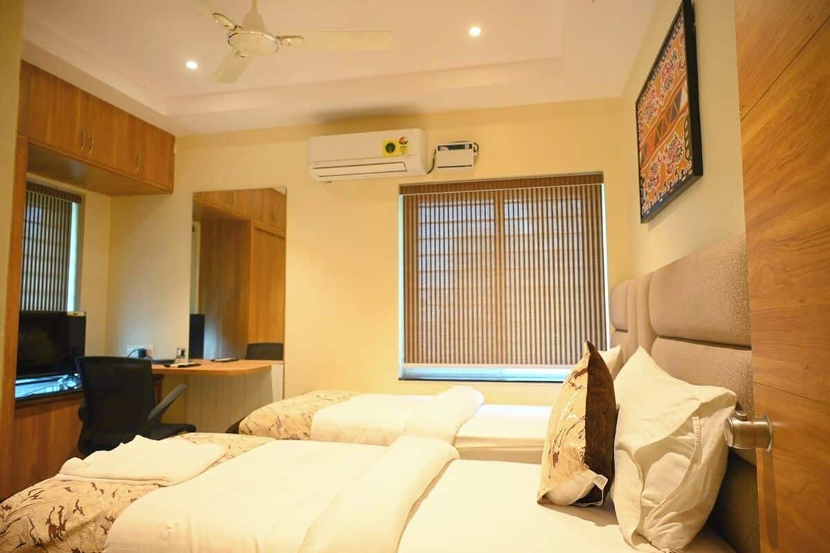 A hotel room with two beds, a desk, a chair, an air conditioning unit, and a window with blinds.