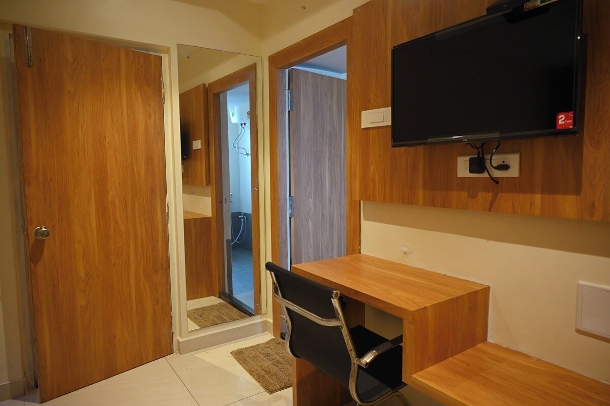 Una habitación compacta con un escritorio de madera, una silla negra, un televisor montado y una puerta que da a otra habitación.