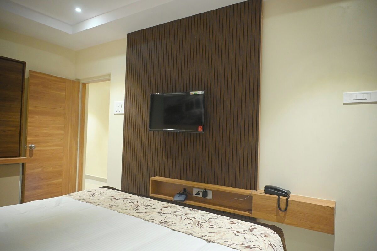 A hotel room with a bed, a TV mounted on a wooden panel wall, a bedside table with a phone and a power outlet, and a wooden door.