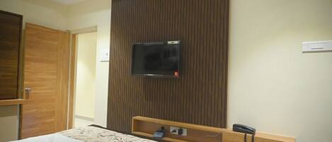 A hotel room with a bed, a TV mounted on a wooden panel wall, a bedside table with a phone and a power outlet, and a wooden door.