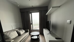 A modern hotel room with a sofa, a small round table, a TV on a stand, and a large window offering a city view. - 1 bed plus condo near JJ market (Bangkok)