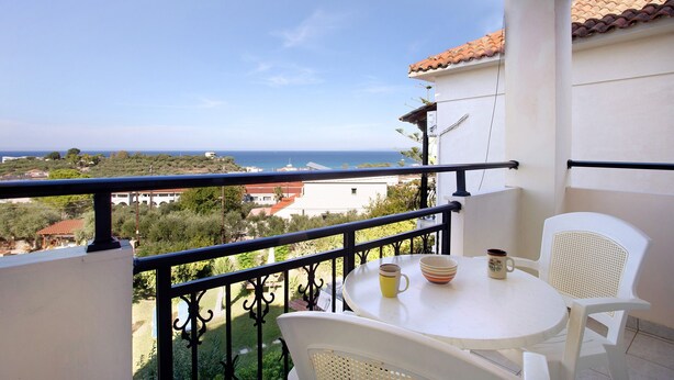 A balcony with a table and chairs, a view of the sea, and a building with a tiled roof. - Studio Apartment "Yiannis 7" with Sea View, Shared Garden & Wi-Fi (Zakynthos)