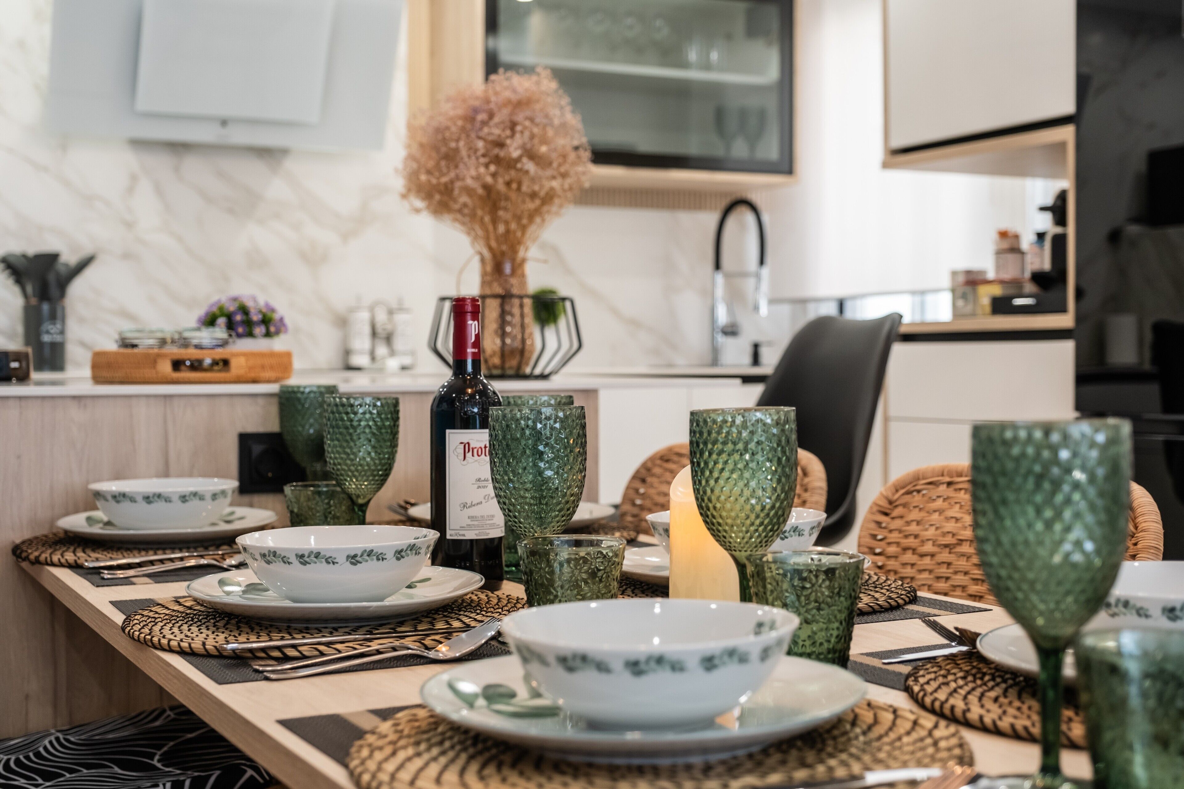 Una mesa de comedor dispuesta con cristalería verde, una botella de vino y una vela.