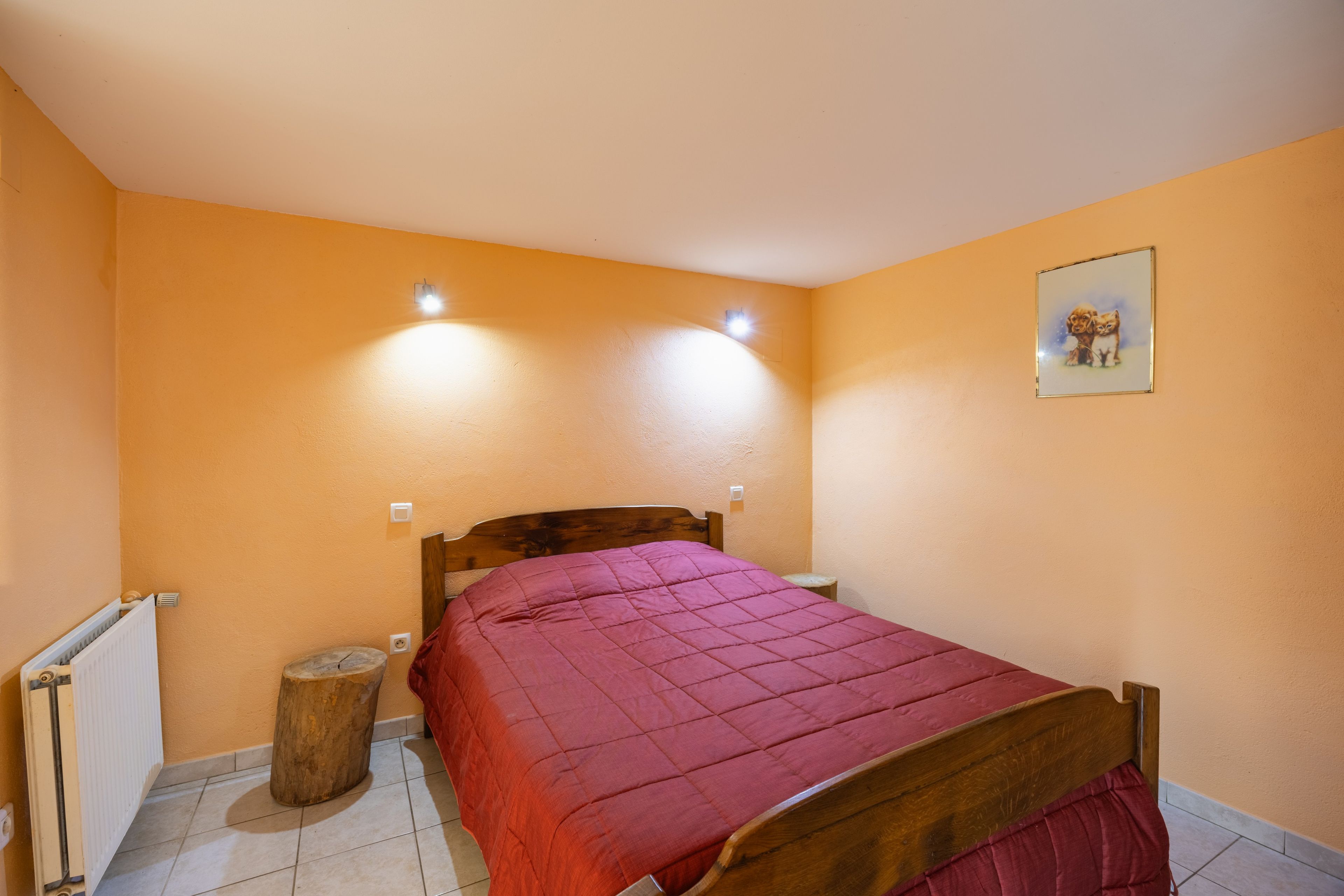 A bedroom with a wooden bed, a red bedspread, a small wooden stool, a radiator, and a wall-mounted light fixture.
