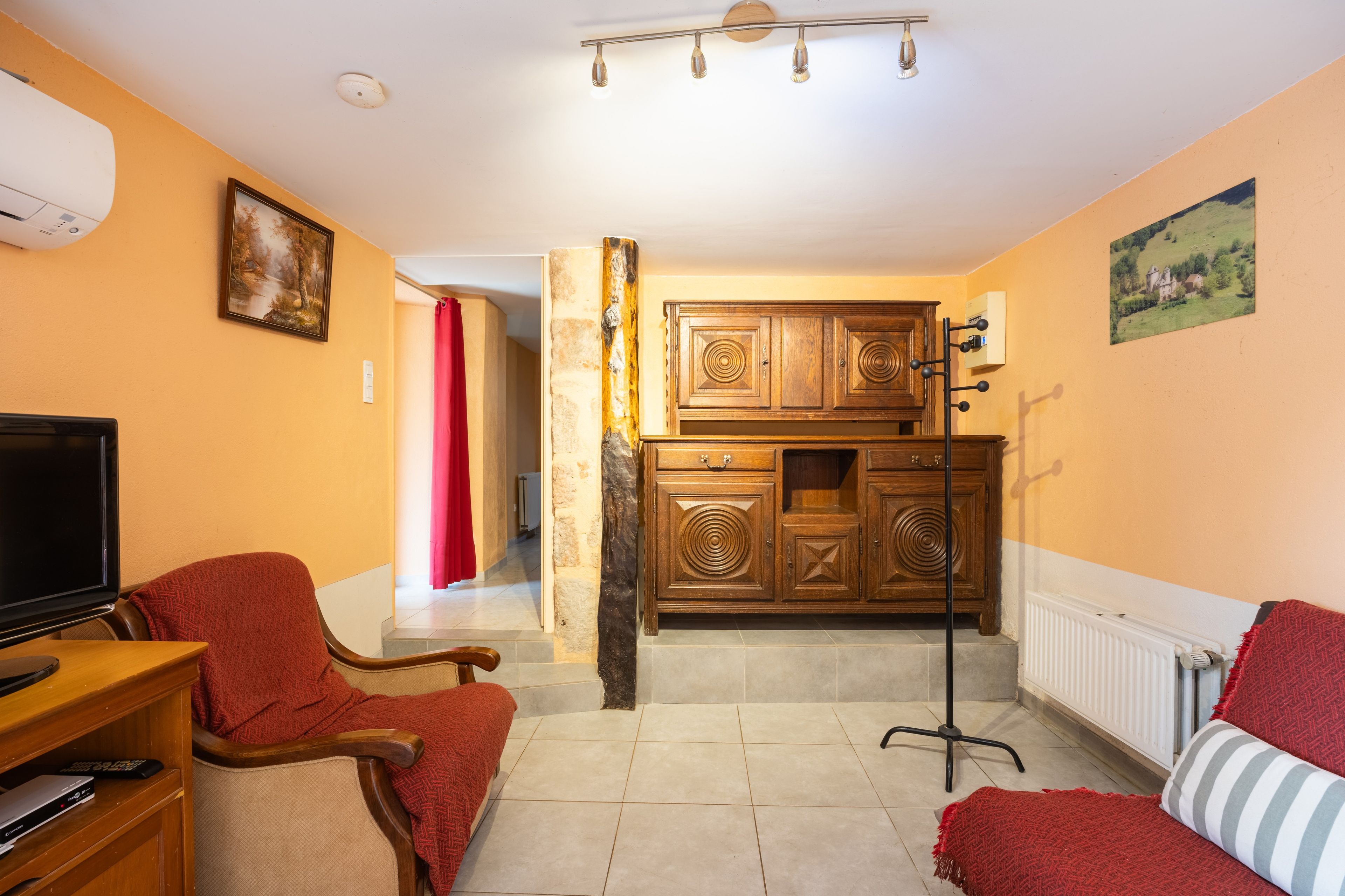 Un salon comprenant un téléviseur, un fauteuil rouge, un canapé, un meuble en bois et un radiateur.