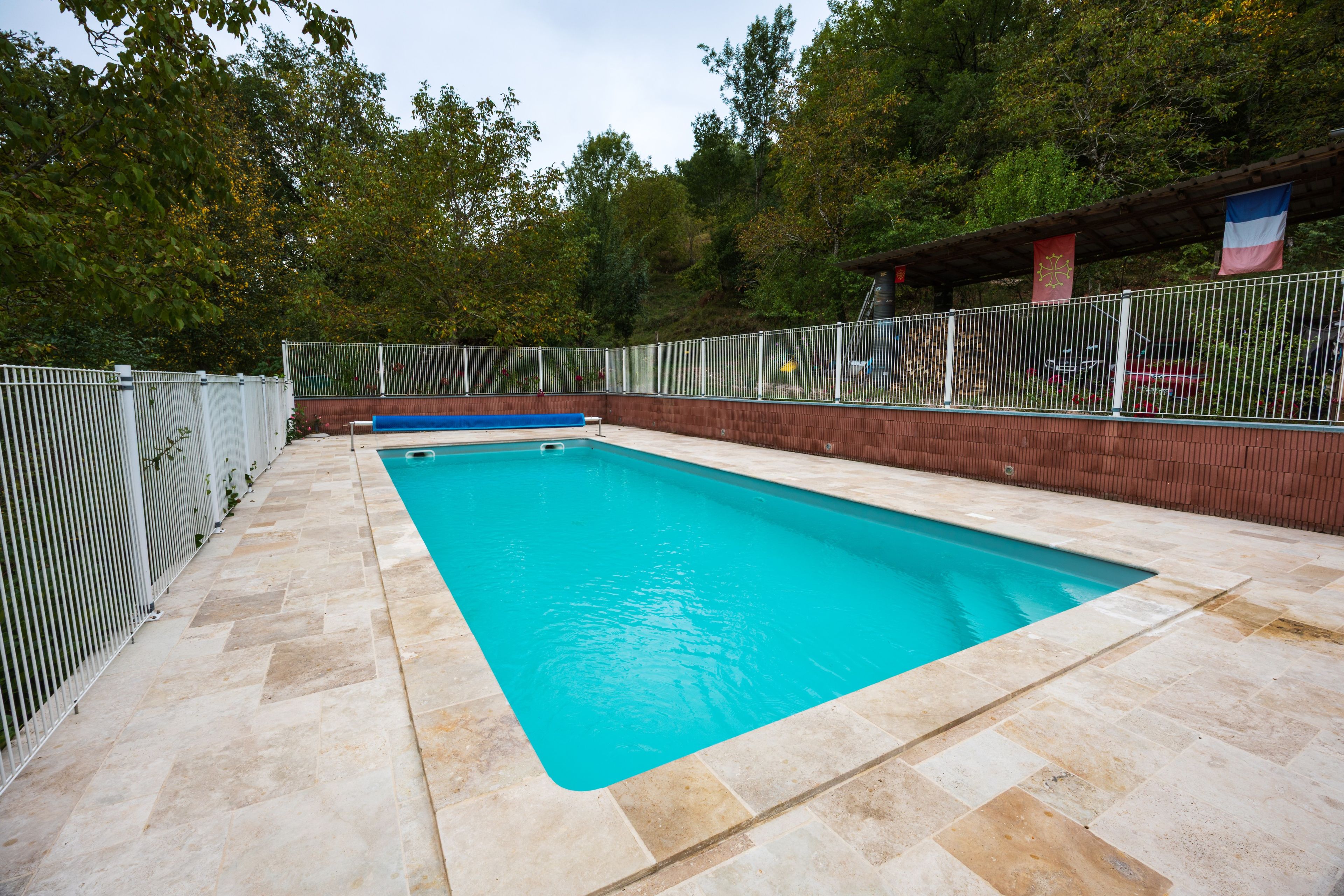 A rectangular swimming pool with a tiled deck, surrounded by a metal fence and trees in the background.