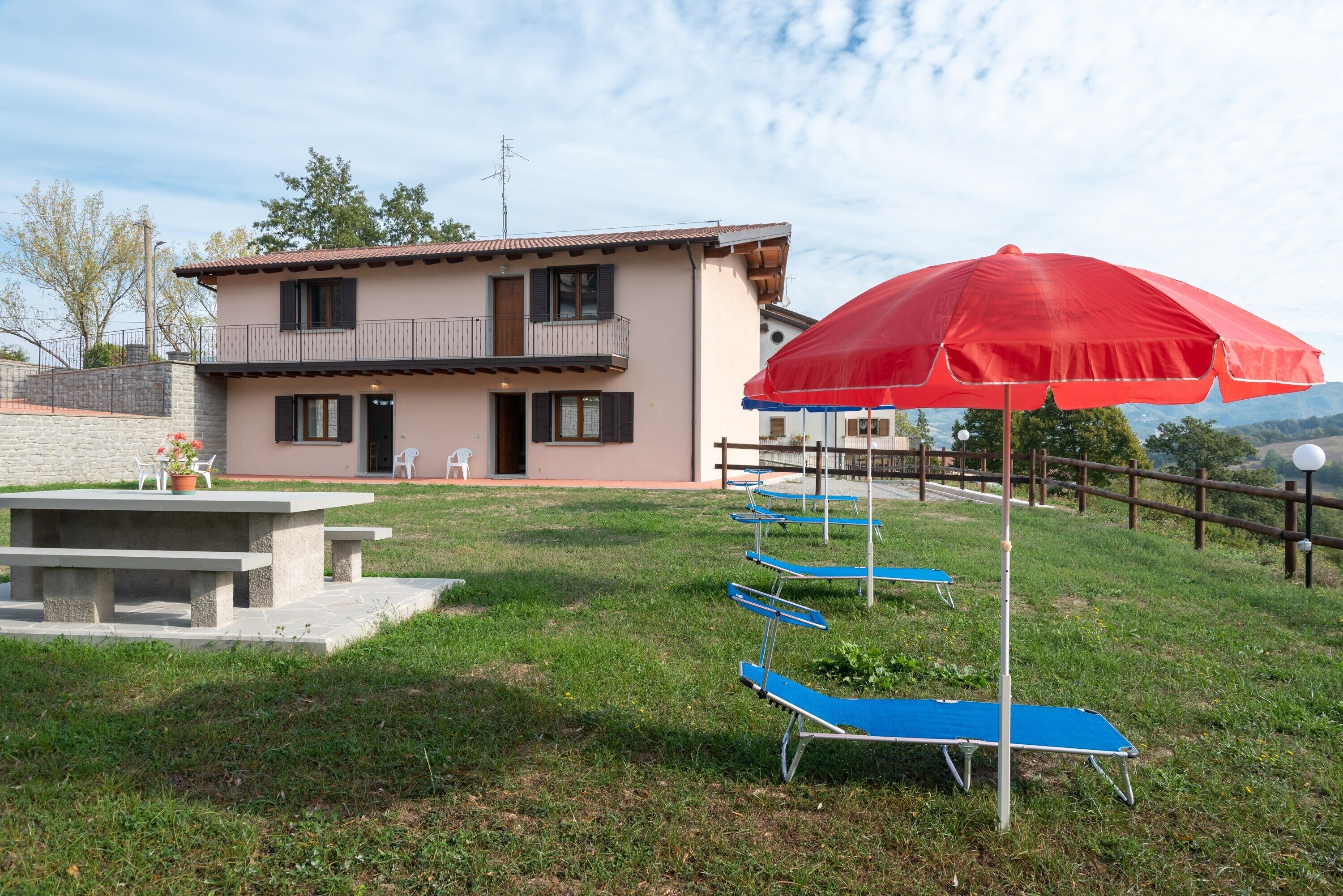 A two-story building with a balcony, a red umbrella, and blue lounge chairs in a grassy area.