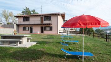 A two-story building with a balcony, a red umbrella, and blue lounge chairs in a grassy area.