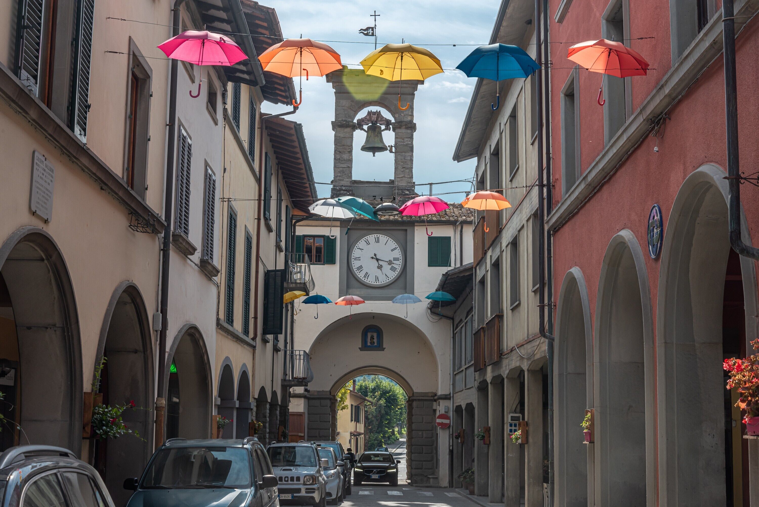 A narrow street with parked cars, a clock tower, and colorful umbrellas.