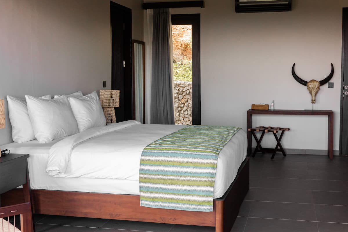 A bedroom with a bed, pillows, a bedside table, a lamp, a curtain, a stone wall, a wooden table, and a bull skull decoration.
