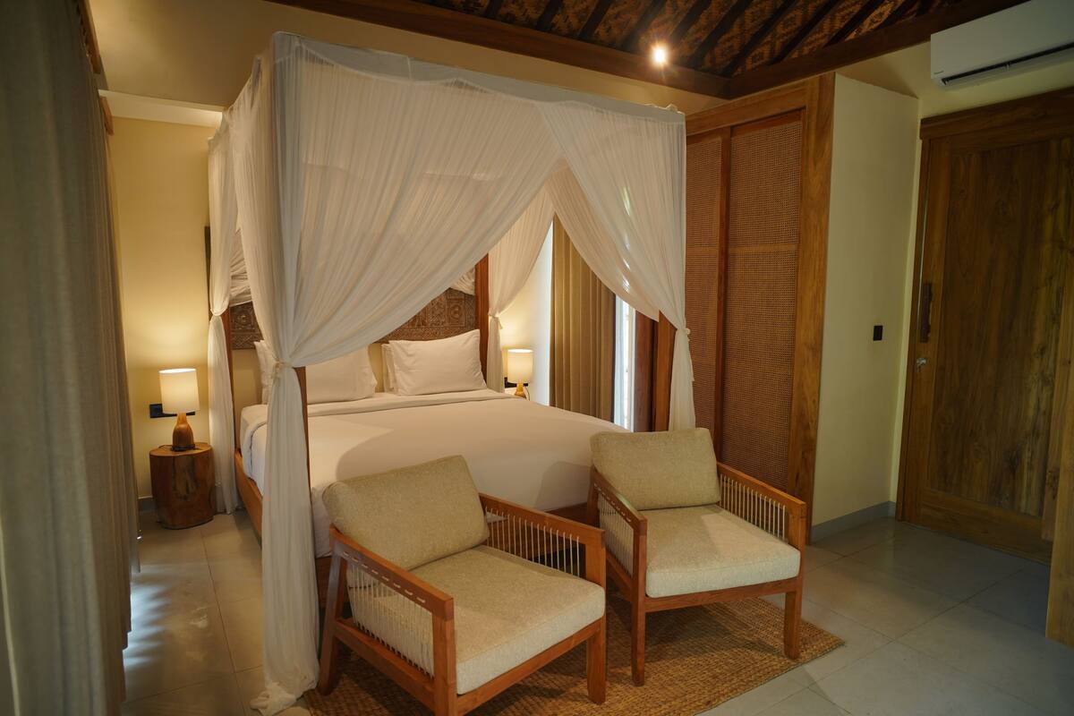 A bedroom with a four-poster bed, two chairs, a nightstand, and a wooden door.