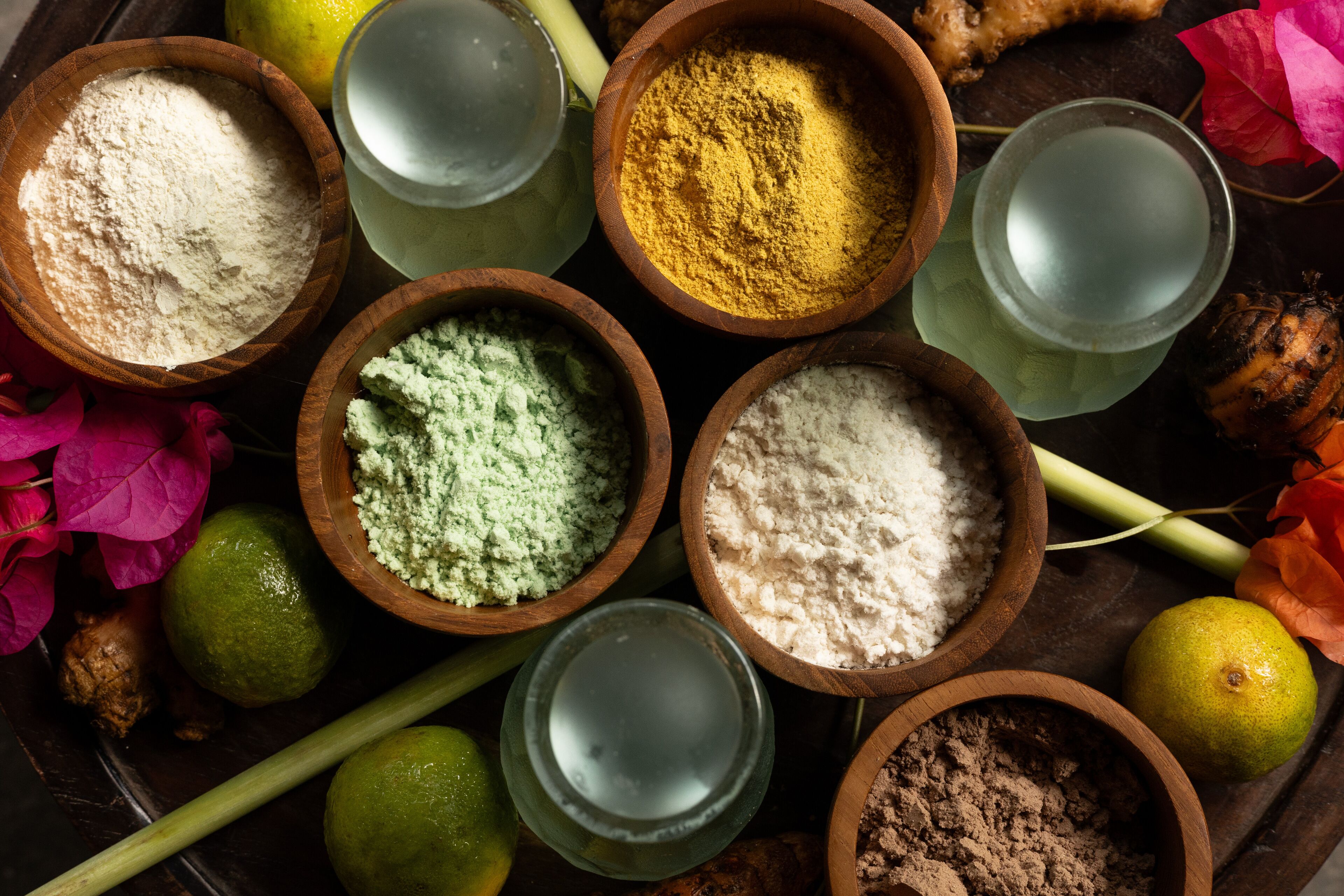 Différents récipients contenant des poudres et des liquides, disposés autour de citrons verts et de fleurs.