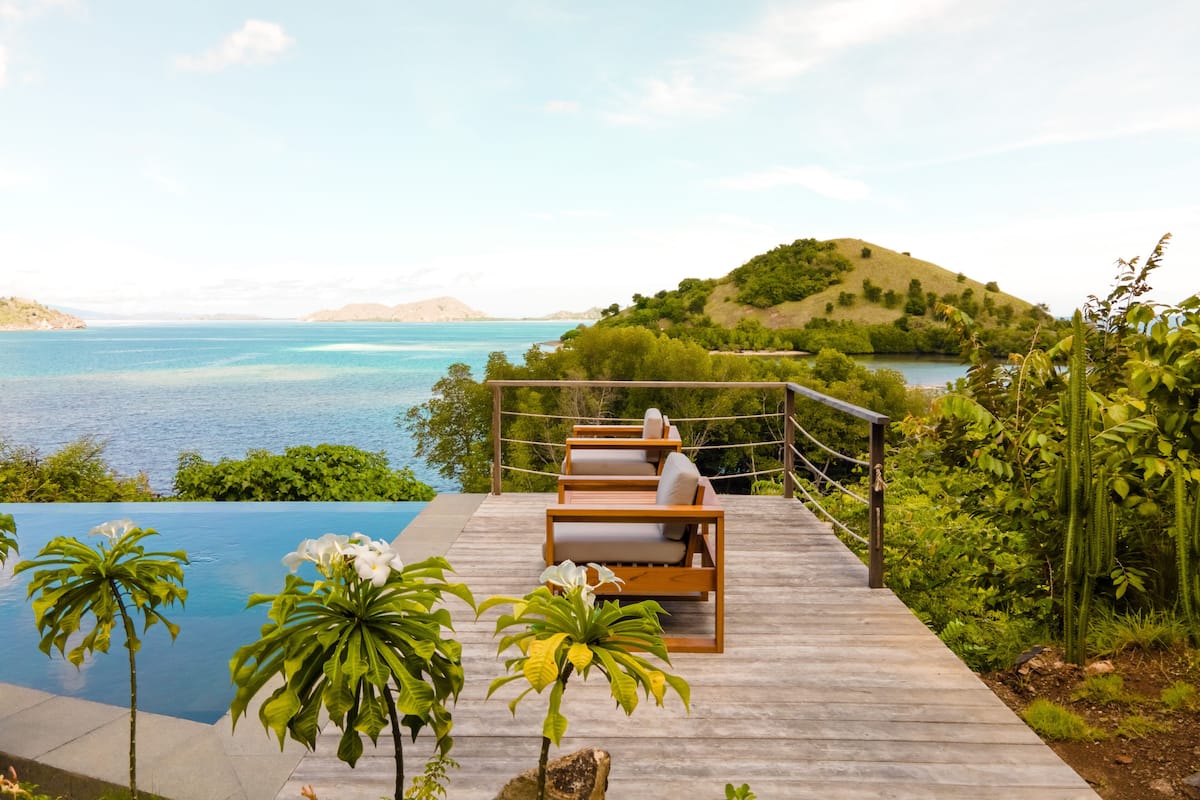 A wooden deck with a pool, overlooking a body of water and a hilly landscape.