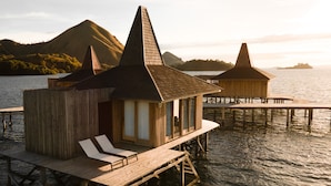 Bungalows sobre el agua con estructuras de madera y sillones de descanso, unidos por una terraza de madera sobre aguas tranquilas, con montañas al fondo.