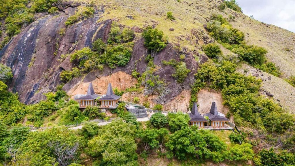 A hillside with three traditional-style buildings with steep roofs, surrounded by lush greenery and rocky terrain.