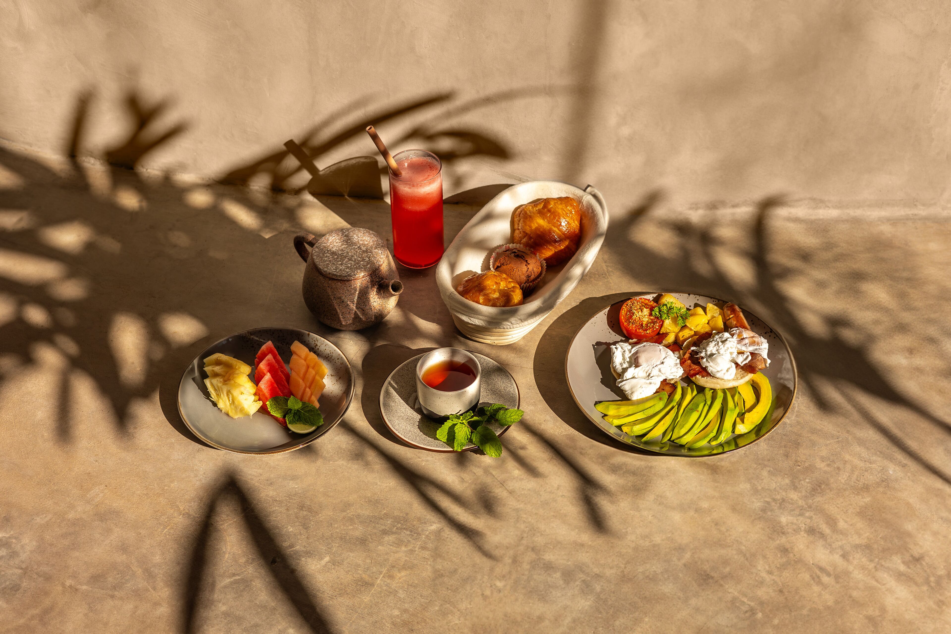 Un tavolo con diversi cibi, tra cui frutta, pasticcini e una bevanda, su una superficie strutturata con ombre proiettate da oggetti vicini.