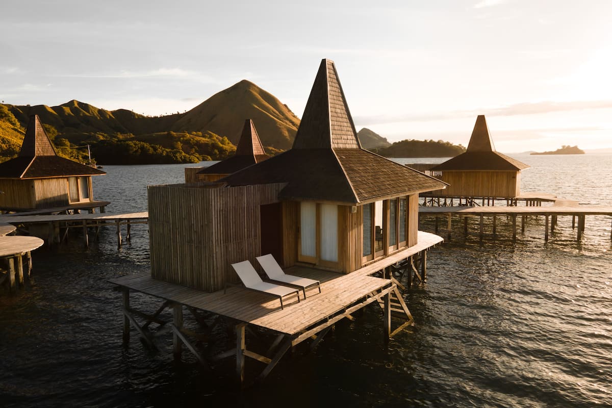 A wooden overwater bungalow with a conical roof, a deck, and a lounge chair.