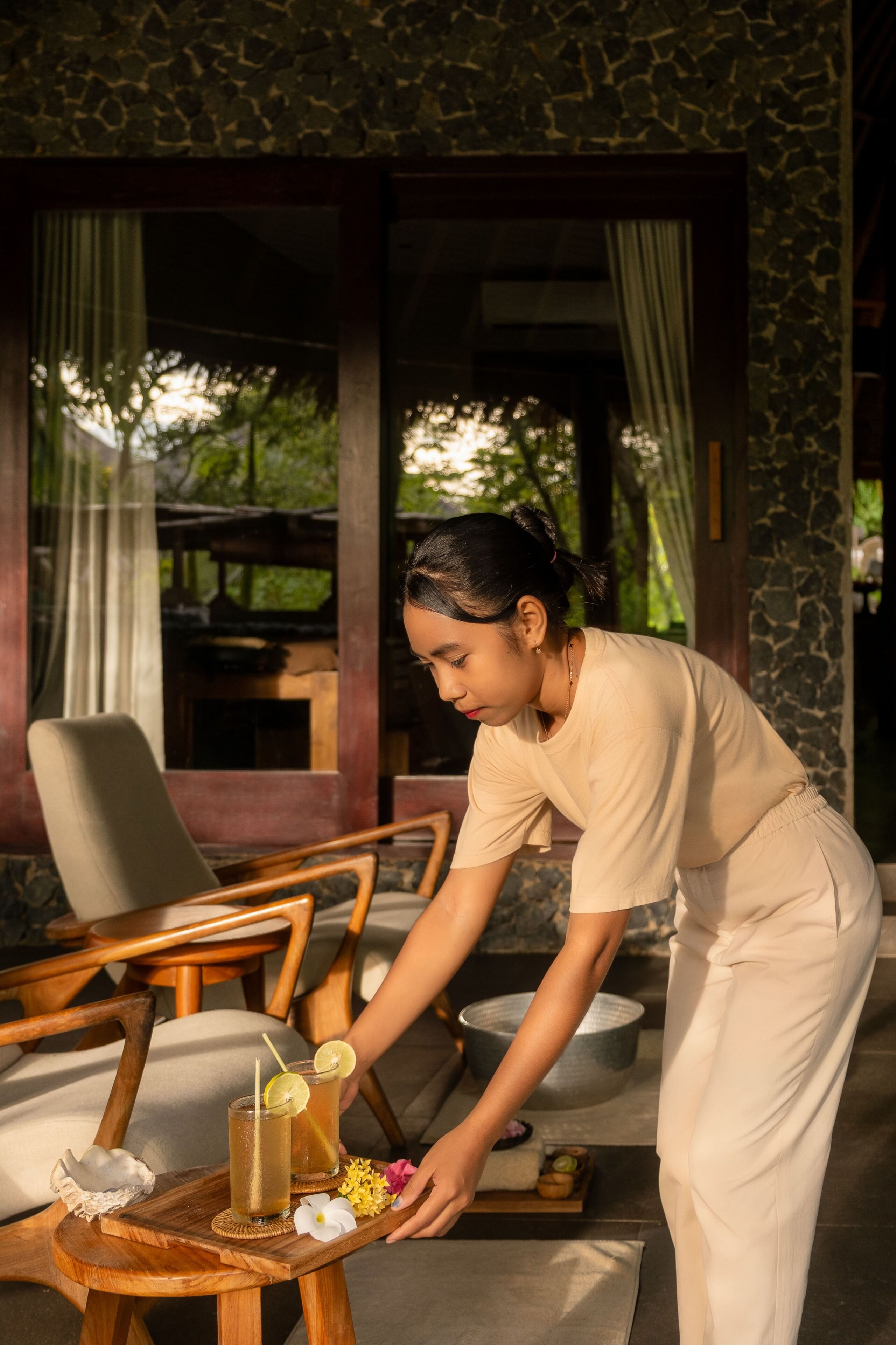 A person serving drinks on a wooden tray with a glass and a bowl, in a room with stone walls and wooden furniture.