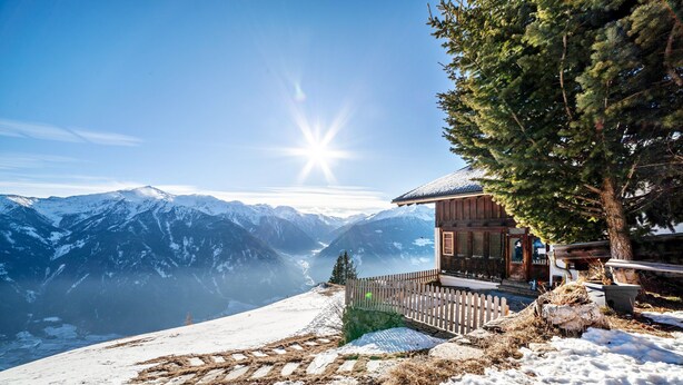 Eine Holzhütte mit einer Holzdiele, umgeben von schneebedeckten Bergen und blauem Himmel.