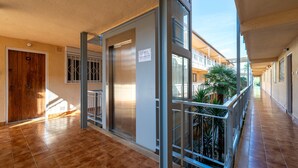Un couloir avec une porte en bois, un ascenseur en métal et un balcon agrémenté de plantes.