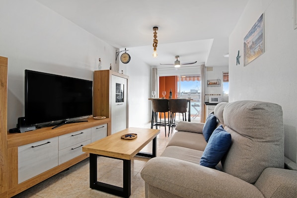 A modern living room with a flat-screen TV, wooden furniture, a sofa with blue cushions, a dining area with chairs, and a view through a large window.