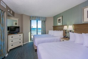 Ein Hotelzimmer mit zwei Betten, einem Fernseher auf einer Kommode, Nachttischen mit Lampen und Blick auf das Meer.