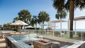 Eine Terrasse direkt am Strand mit einer langen, gestreiften Couch, einer Feuerstelle aus Stein und Gartenmöbeln.