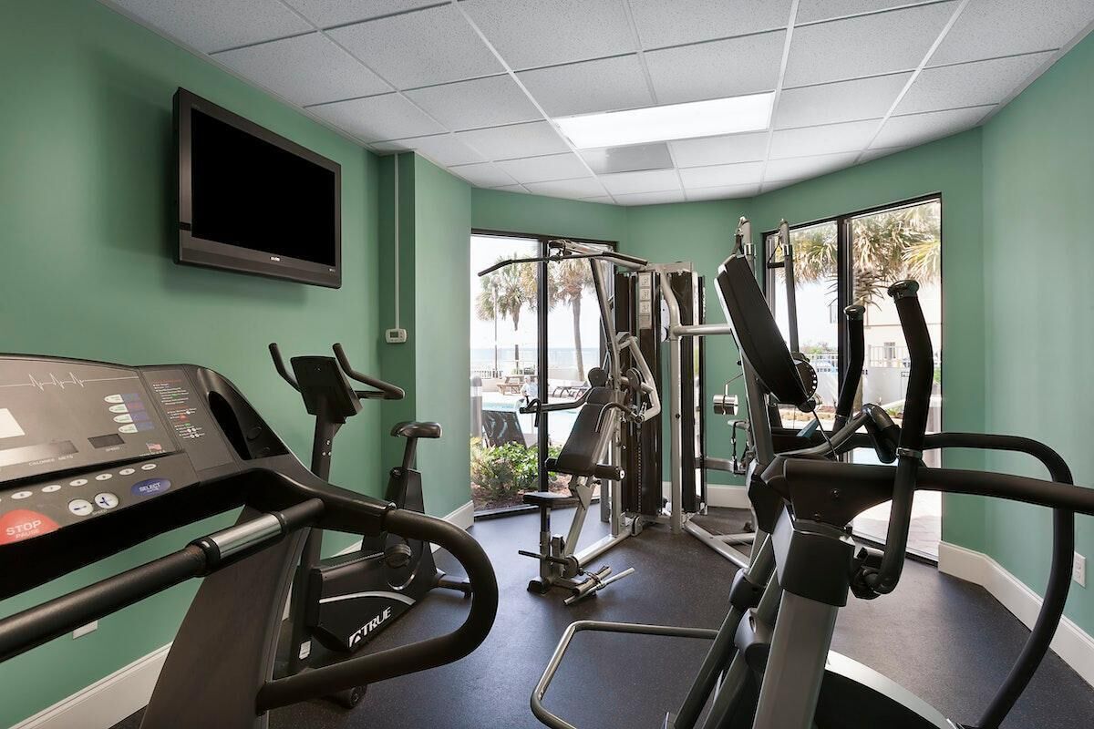 Een fitnessruimte met cardioapparatuur, een televisie en grote ramen met uitzicht op palmbomen.