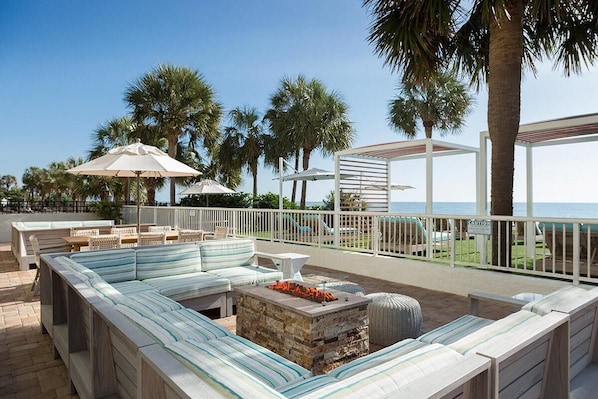 A beachside patio with a long, striped sofa, a stone fire pit, and outdoor seating.