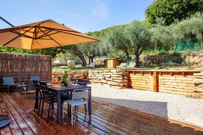 Un patio exterior con una terraza de madera, una mesa con sillas, un gran paraguas y un muro de piedra.