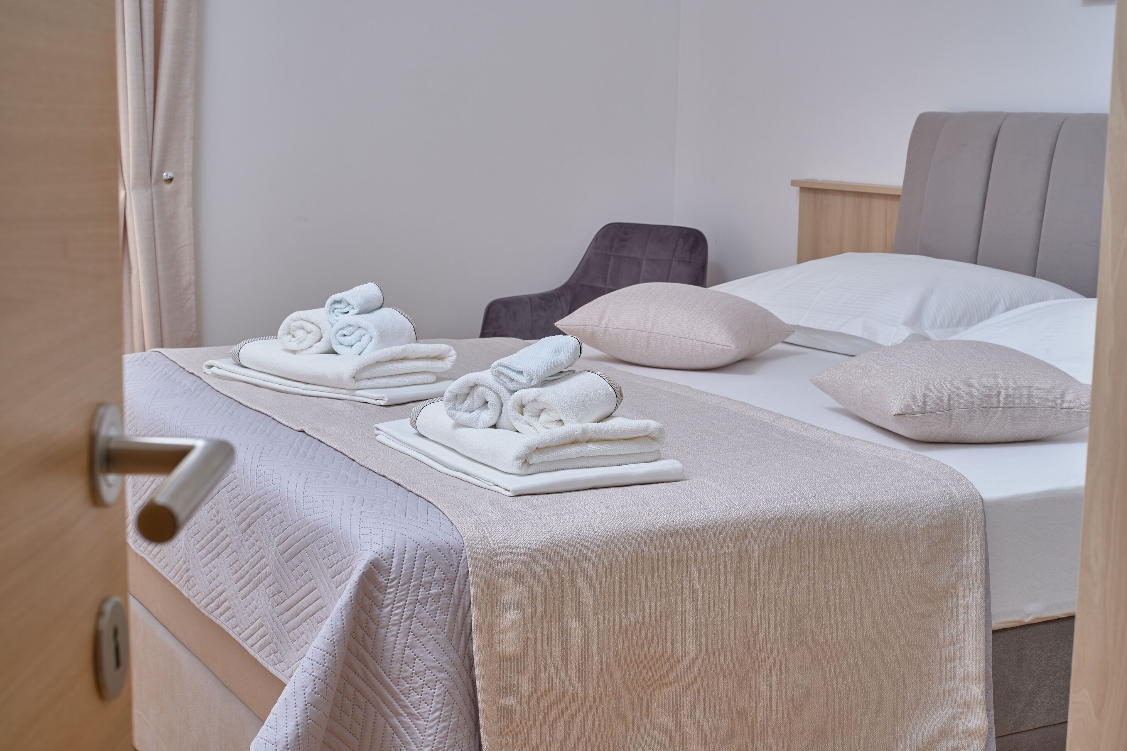 Een net opgemaakt bed met een beige sprei, kussens en opgerolde handdoeken op een houten nachtkastje.