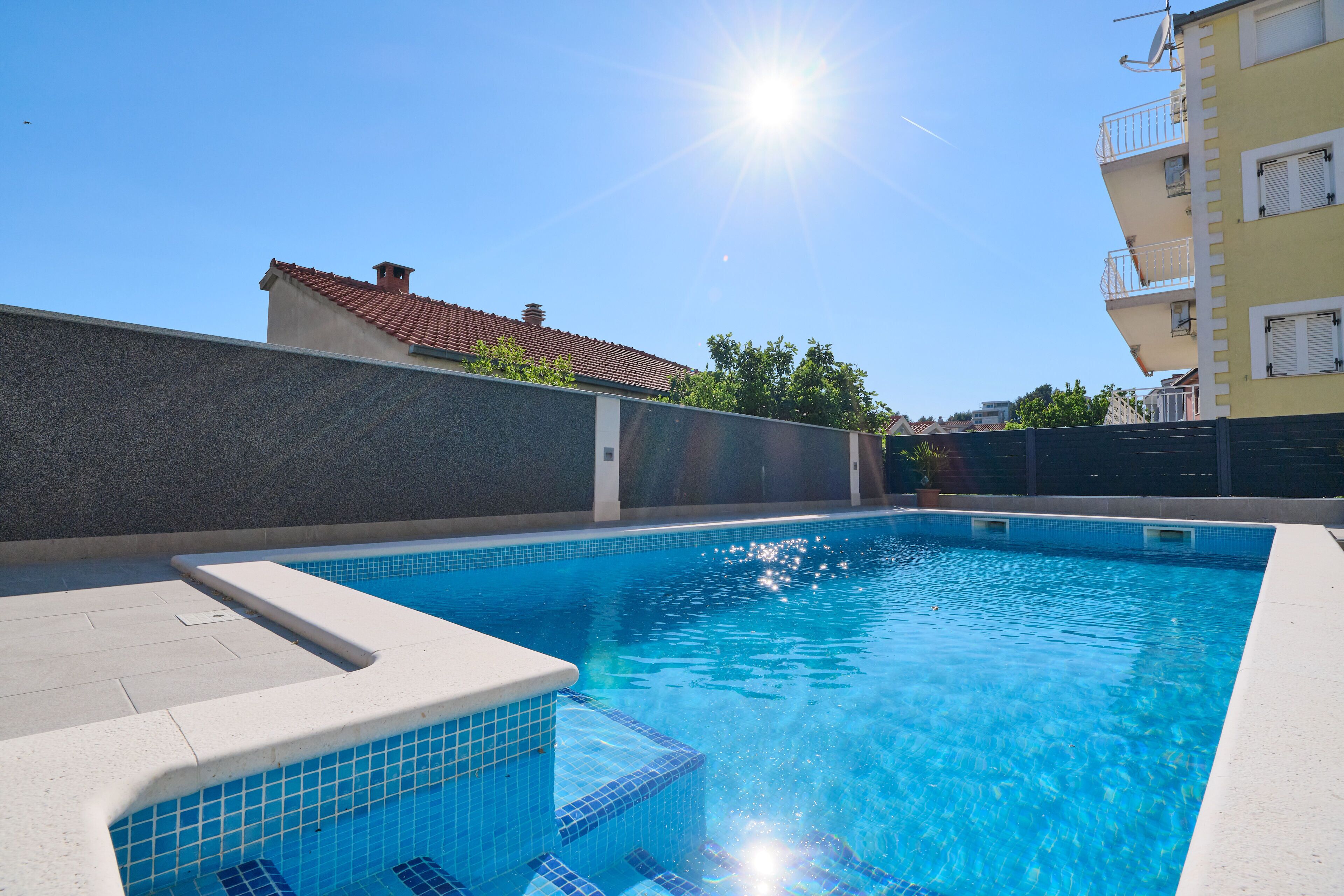A swimming pool with clear blue water, surrounded by a stone wall and a building with balconies.