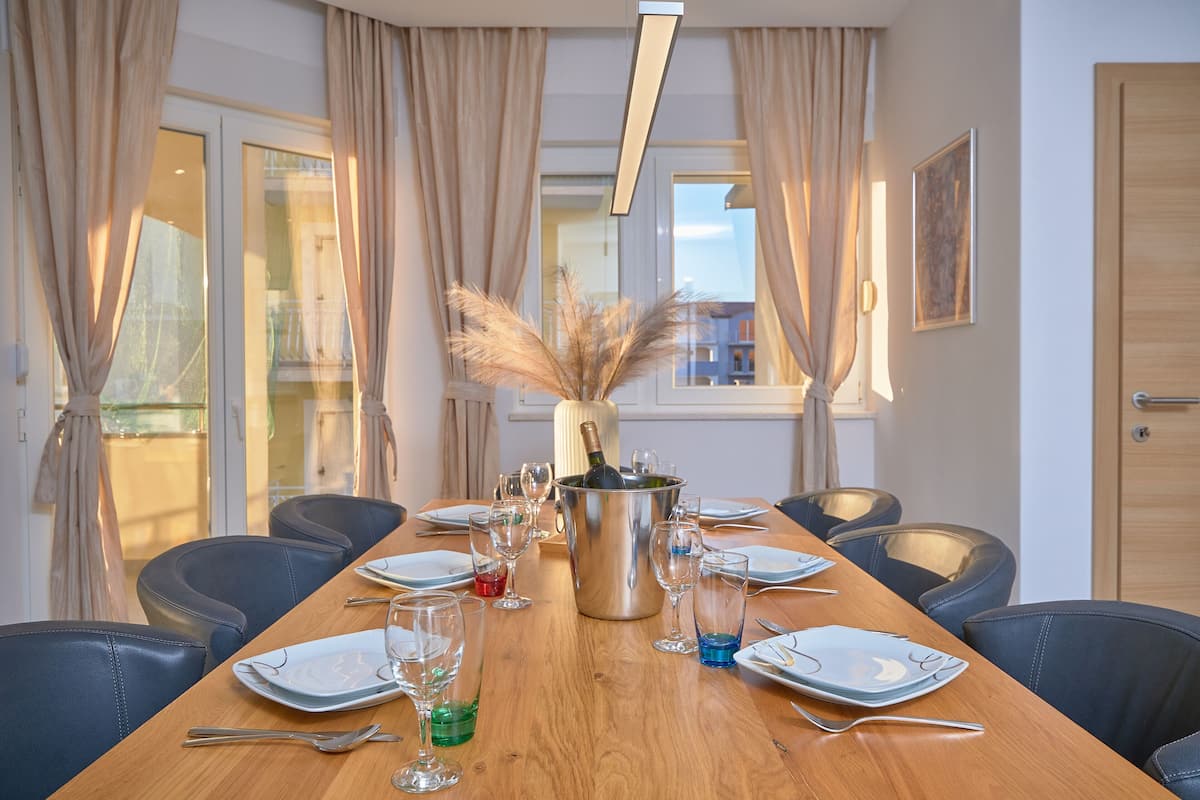 A dining area with a long wooden table set for a meal, surrounded by chairs, with a bottle of champagne in the center.