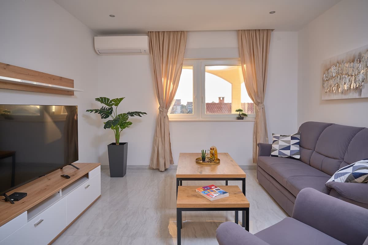 A modern living room with a flat-screen TV, a wooden coffee table, a grey sofa, and a large window with curtains.